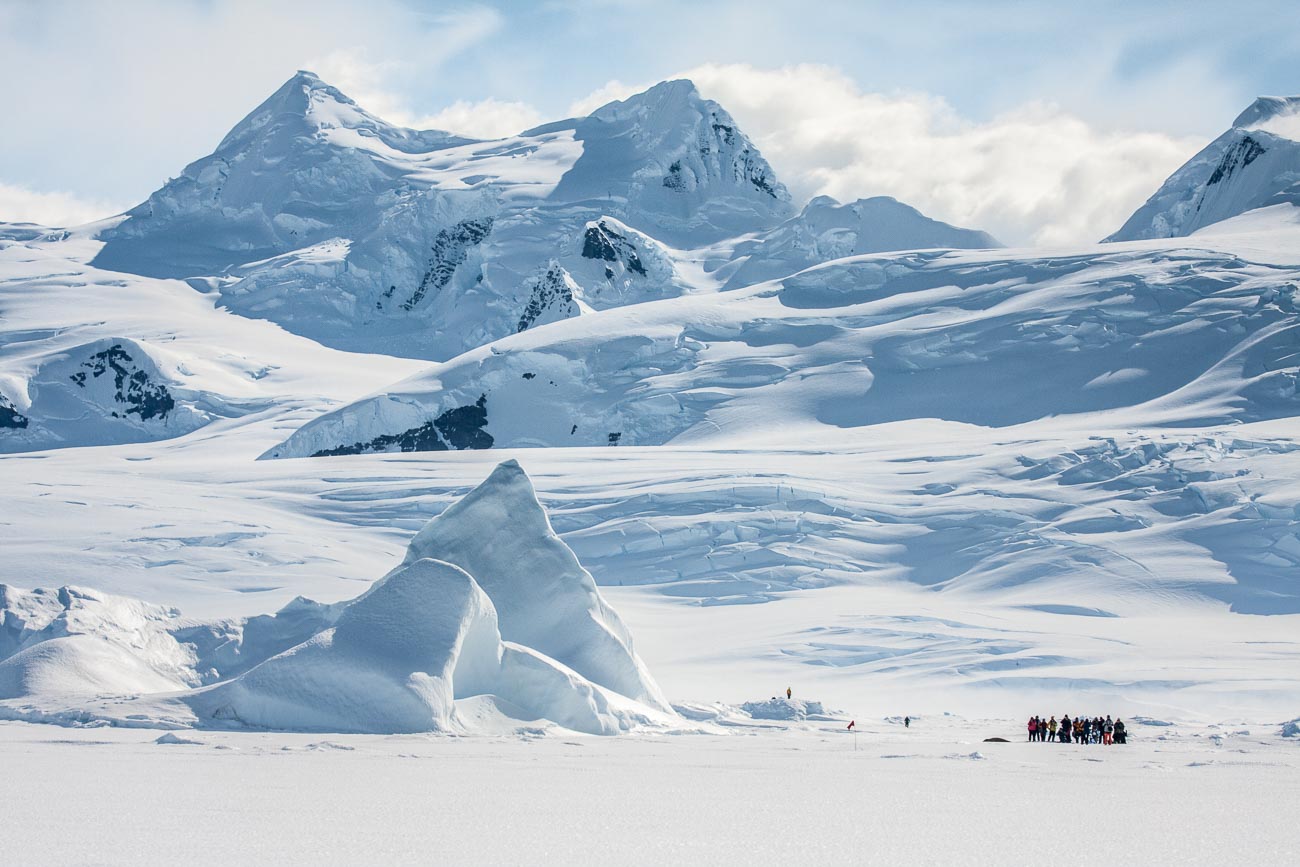 A group of tourists walking on the fast ice in Lallemand bay, watching a seal, surrounded by a grounded iceberg with glaciers in the background